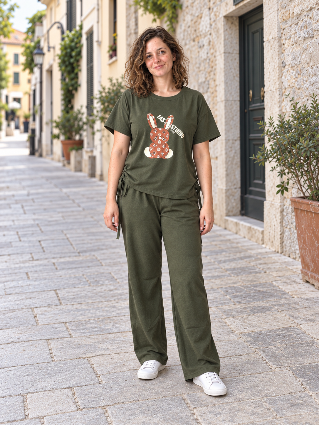 Model wearing an olive-green rabbit graphic top and matching wide-leg trousers on a sunlit street.